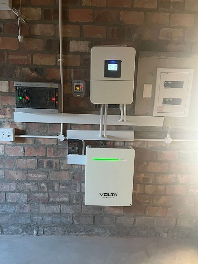 The photo shows a solar energy installation mounted on a brick wall. The setup includes:  1. A *Volta Stage 1* battery unit (bottom) with a green status indicator. 2. An inverter (top) with a digital display, likely managing energy flow between the battery, grid, and load. 3. An *INGE 5kW AC Box* (left) with green and red LEDs indicating operational status and safety warnings. 4. Associated distribution panels and wiring for integrating the system with the electrical grid.  The installation appears to be a hybrid solar power system for energy storage and management. 🔋👍